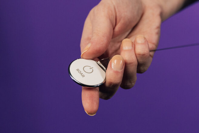 Close-up of a healthcare worker in dark scrubs holding a white ROAR button clipped to a retractable badge reel, against a purple background.