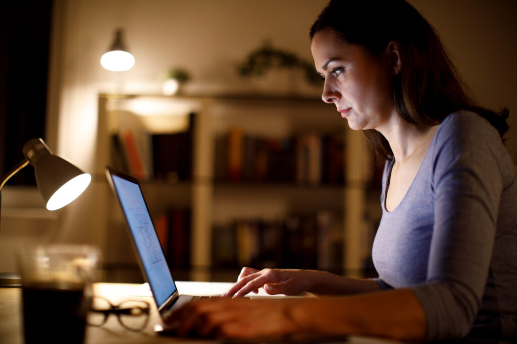 woman working alone at night on laptop with bookshelf in background
