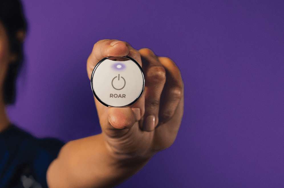 Hand holding up a round white ROAR panic button with an illuminated purple indicator light, against a purple background.